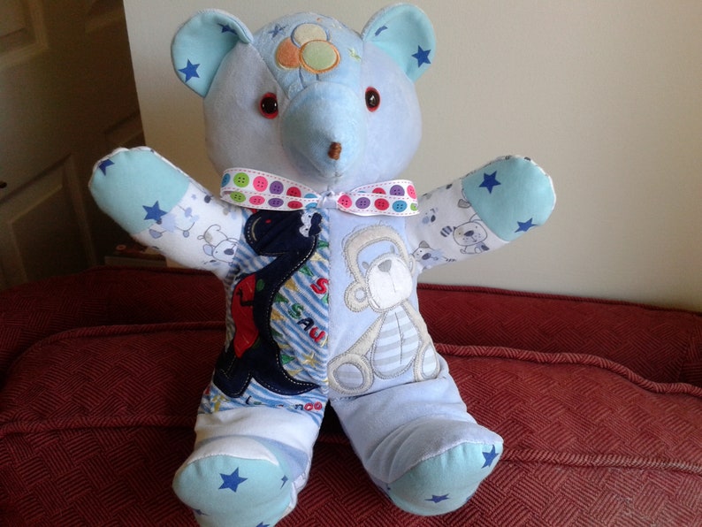 Op de afbeelding: Een blauwe en witte patchwork teddybeer met een rode knoopneus en een lint met kleurrijke knopen om zijn nek. De beer zit op een rode bank.