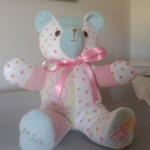 Op de afbeelding: Een handgemaakte patchwork teddybeer met een roze lint om zijn nek. De beer is gemaakt van verschillende stoffen, waaronder stippen, bloemen en effen kleuren. De poten van de beer zijn blauw en de beer zit op een grijs oppervlak. De beer heeft de naam "Mia" geborduurd op een van zijn poten.