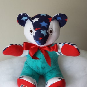 Op de afbeelding: Een handgemaakte pluche teddybeer met een blauw, wit en rood sterrenpatroon op zijn hoofd en armen. De beer draagt een turquoise outfit met een rode strik en heeft de woorden "Arsenal" en "Sh" op zijn voeten.