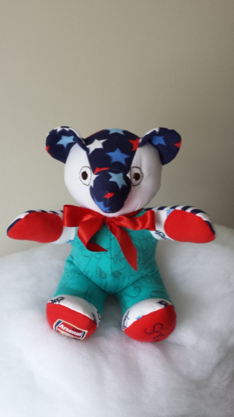 Op de afbeelding: Een handgemaakte pluche teddybeer met een blauw, wit en rood sterrenpatroon op zijn hoofd en armen. De beer draagt een turquoise outfit met een rode strik en heeft de woorden "Arsenal" en "Sh" op zijn voeten.