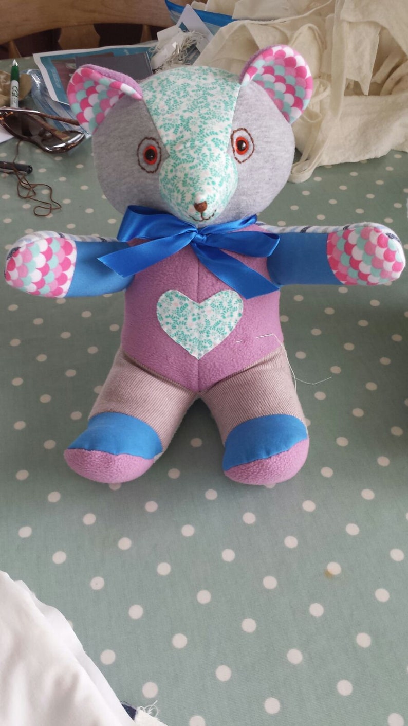 Op de afbeelding: Een handgemaakte patchwork teddybeer met een roze, blauwe en witte bloemenprint. De beer heeft een blauw lint om zijn nek en een hartvormige patch op zijn borst.