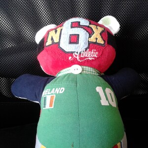 Op de afbeelding: Een pluche teddybeer die een groen-wit shirt draagt met het woord "IRELAND" en het nummer "10" op de rug. De beer draagt ook een rood shirt met de letters "N6X" en het woord "Athletic" op de voorkant.