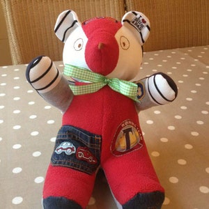 Op de afbeelding: Een rode en witte patchwork teddybeer met een groene strik, blauwe jeans en verschillende geborduurde patches, waaronder "Ik hou van mama en papa" en "Winnend team".