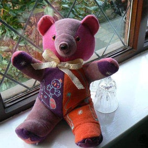 Op de afbeelding: Een patchwork teddybeer van paarse, roze, oranje en witte stof. De beer heeft een strik om zijn nek en zit op een wit oppervlak.