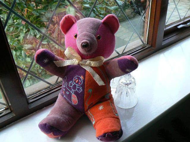 Op de afbeelding: Een patchwork teddybeer van paarse, roze, oranje en witte stof. De beer heeft een strik om zijn nek en zit op een wit oppervlak.