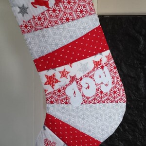 Peut inclure: Une chaussette de Noël matelassée avec des motifs rouges, blancs et gris. Elle présente des motifs de rennes et d'étoiles, avec le mot "joy" appliqué. Le haut et le bas sont rouges.