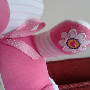 Op de afbeelding: Roze en wit gestreepte teddybeer van stof met een roze strik met witte stippen. De beer heeft een roze bloem geborduurd op een arm.