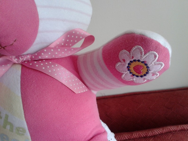 Op de afbeelding: Roze en wit gestreepte teddybeer van stof met een roze strik met witte stippen. De beer heeft een roze bloem geborduurd op een arm.