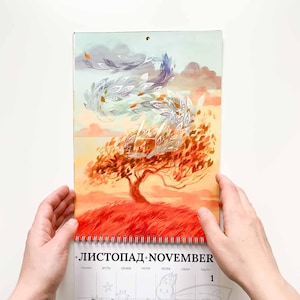 Op de afbeelding: Een kalender met een illustratie van een herfsttafereel met een boom en wervelende bladeren. De kalender is open voor november, met zichtbare data. Het kunstwerk heeft warme kleuren en de kalender wordt door twee handen vastgehouden.