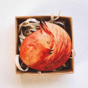 May include: A wooden fox figurine in a curled sleeping position, painted in shades of orange and red, rests inside a small, square cardboard box. The fox has a black-tipped ear and a closed-eye design. The box is filled with shredded paper.