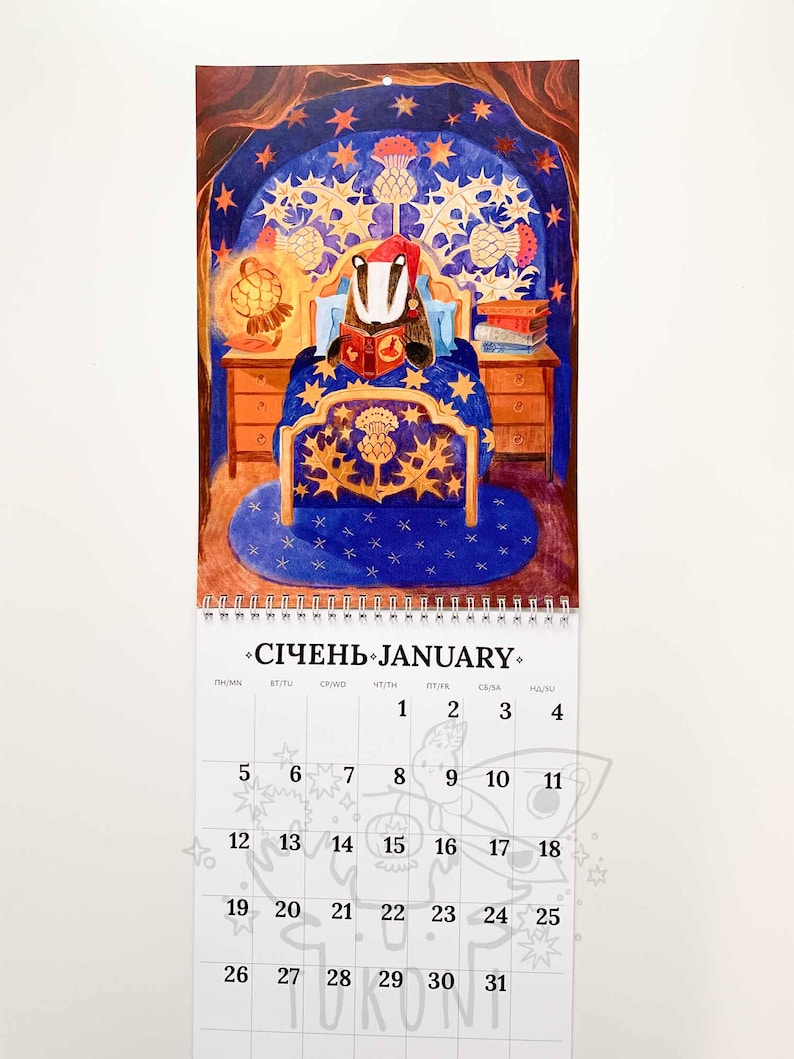 Op de afbeelding: Een wandkalender voor januari met een illustratie van een das die in bed leest, omringd door sterren en distels. De kalender toont de data van de maand, van 1 tot en met 31.