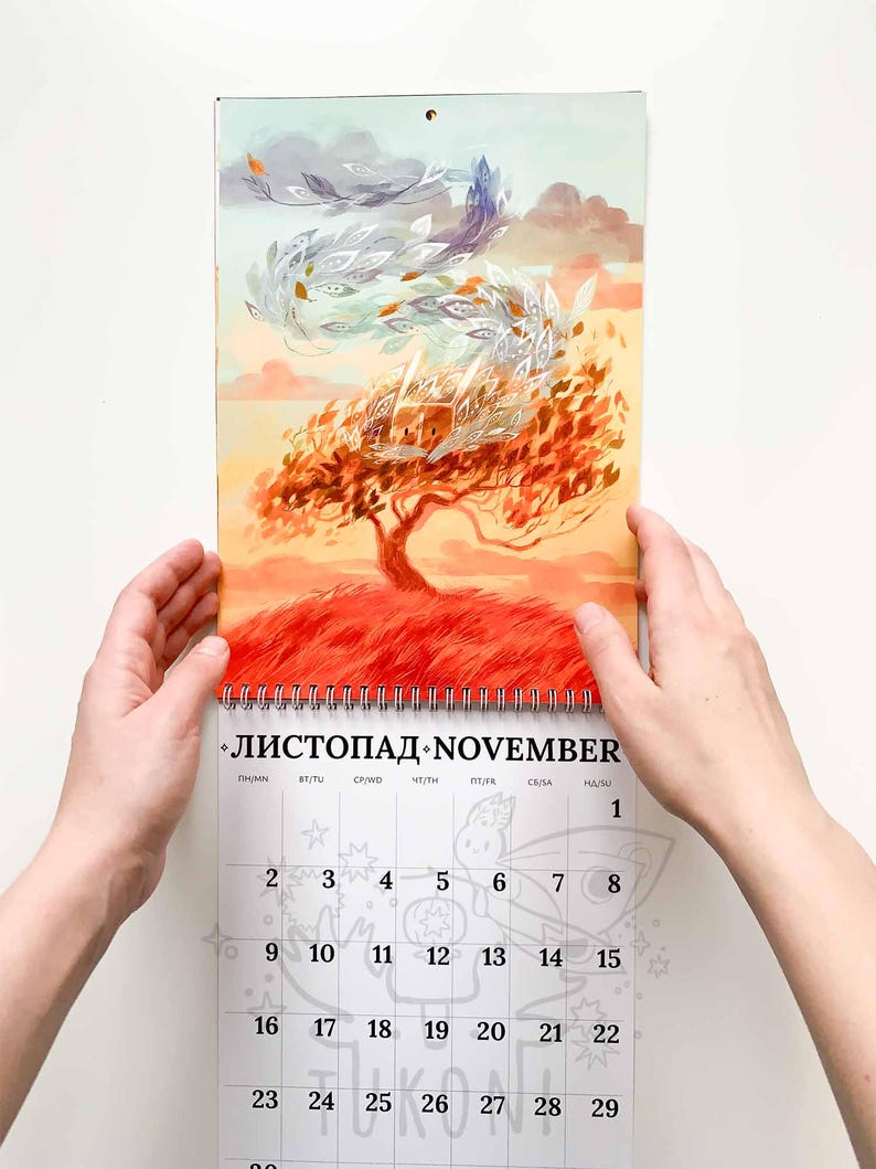 Op de afbeelding: Een kalender met een illustratie van een herfsttafereel met een boom en wervelende bladeren. De kalender is open voor november, met zichtbare data. Het kunstwerk heeft warme kleuren en de kalender wordt door twee handen vastgehouden.