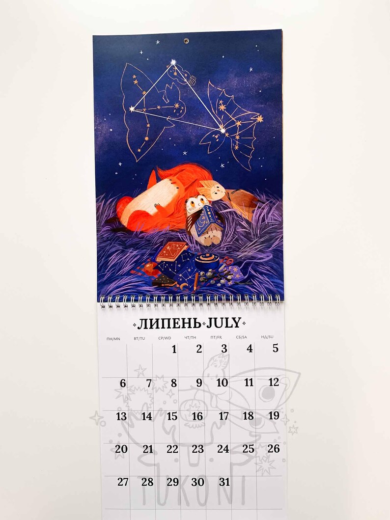 Op de afbeelding: Een juli-kalender met een fantasie-illustratie van een vos, uilen en een theepot onder een sterrenhemel. De kalender heeft een spiraalbinding en een maandelijks overzicht met data en het woord "juli".