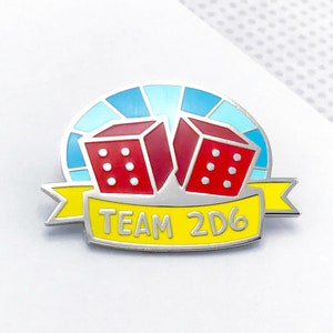 May include: A silver-colored enamel pin featuring two red dice with white dots, set against a blue and light blue background. A yellow banner below reads "TEAM 2D6" in white letters. The pin is likely a collectible or accessory.