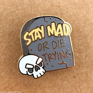 May include: A gray tombstone-shaped enamel pin with the text "STAY MAD OR DIE TRYING" in yellow. A white skull with black eyes and a gold border is on the left side. The pin has a gold border and the text "OMGCHANNERS" at the bottom.