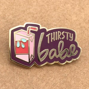 May include: A pin with a purple border and the words "Thirsty Babe" in gold script. The pin features a pink juice box with a red straw and blue water droplets. The pin is on a brown surface.
