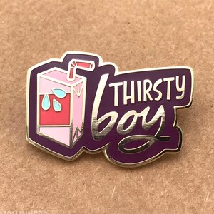 May include: A pin featuring a juice carton design and the text "THIRSTY BOY". The juice carton is pink and red with blue water droplets. The pin has a gold border and is set against a brown background.
