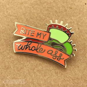 May include: A gold-colored enamel pin featuring a Venus flytrap design. The flytrap is green with red and brown accents. A banner across the pin reads "BITE MY whole ass" in silver lettering. The pin is set against a neutral background.
