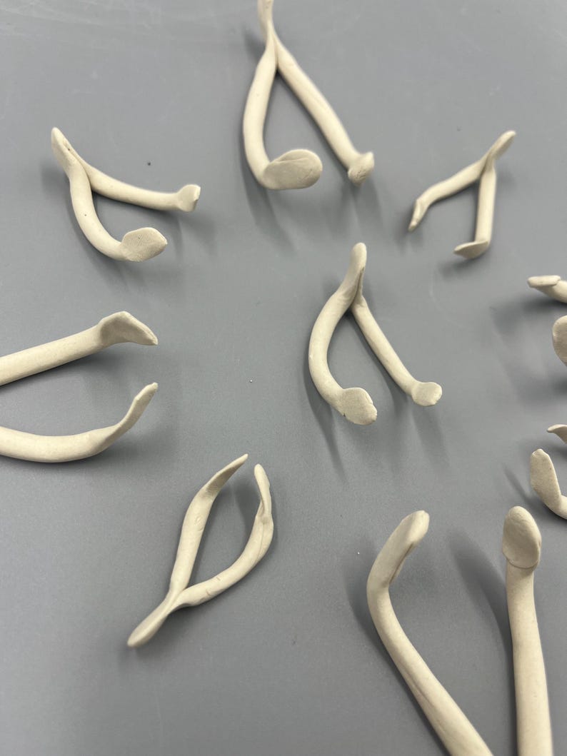 May include: A collection of small, off-white ceramic wishbones scattered on a gray surface. The wishbones have a smooth, curved design with rounded ends, suggesting a handmade craft or decorative item. The arrangement is random, with some wishbones overlapping.