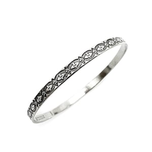 May include: A silver bangle bracelet with an intricate floral pattern. The bracelet is made of a shiny, metallic material and features a repeating design of stylized flowers and leaves. The design is dark against the silver background.