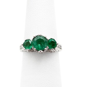 May include: A silver ring with three emerald green gemstones set in a delicate, ornate design.