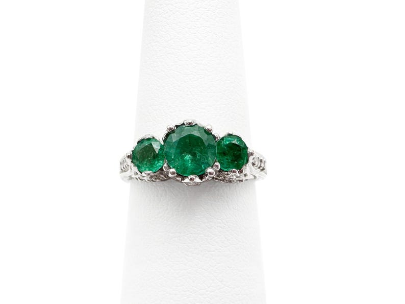 May include: A silver ring with three emerald green gemstones set in a delicate, ornate design.