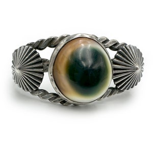 May include: A silver bracelet with a green and white abalone shell inlay. The bracelet has two silver sunburst designs on either side of the shell.