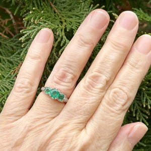 May include: A silver ring with three emerald green gemstones set in a prong setting. The ring is on a person's finger.