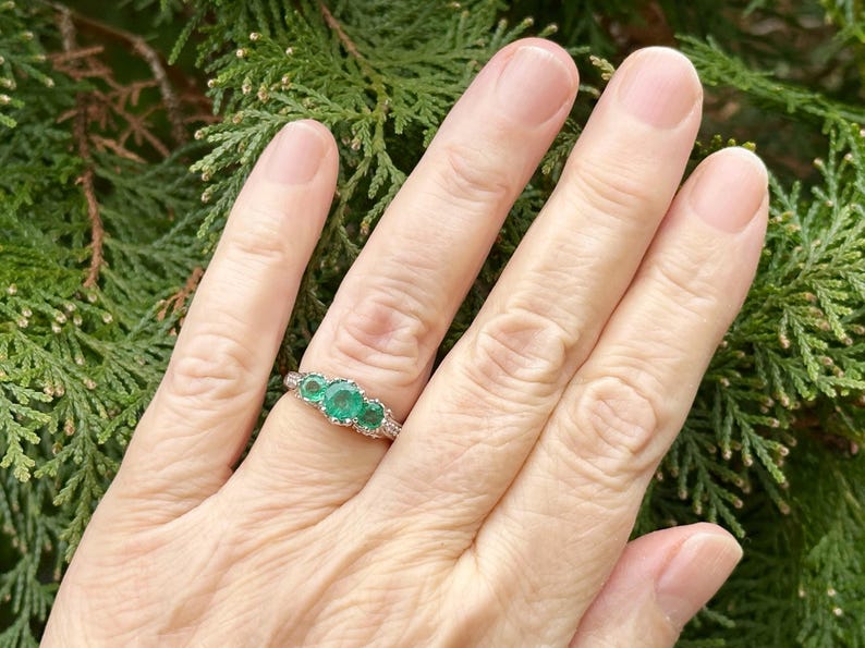 May include: A silver ring with three emerald green gemstones set in a prong setting. The ring is on a person's finger.