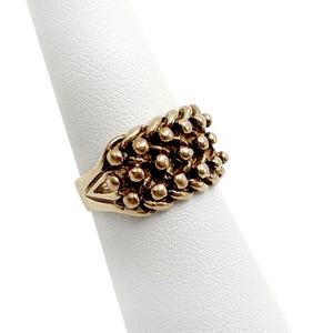 K&ouml;nnte beinhalten: Ein goldener Ring mit einem strukturierten Design aus kleinen, runden Perlen, die in einem gewebten Muster angeordnet sind.