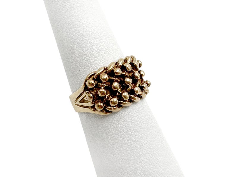 K&ouml;nnte beinhalten: Ein goldener Ring mit einem strukturierten Design aus kleinen, runden Perlen, die in einem gewebten Muster angeordnet sind.