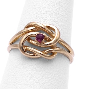May include: A gold ring with a knot design and a small, red gemstone in the center.