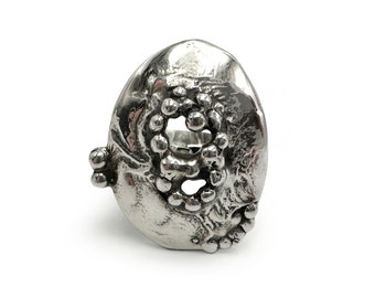 May include: A silver ring with a textured, organic shape and a circular opening. The ring is adorned with small, raised dots around the opening.