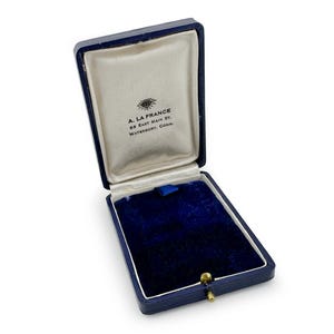 May include: Open blue jewelry box with a velvet interior and a cream-colored satin lining. The box is rectangular with a gold-colored clasp. The text "A. LA FRANCE" is printed on the inside of the lid.