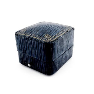 Op de afbeelding: Een kleine, vierkante, marineblauwe sieradendoos met een getextureerd oppervlak. Het deksel is versierd met een decoratieve gouden rand en een bladachtig ontwerp. De doos heeft een zilverkleurige knoopsluiting.