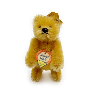 May include: A small, golden-brown teddy bear with black eyes and a button nose. The bear is wearing a round tag that says "Original Teddy" with a smaller teddy bear image inside.