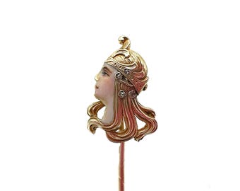 May include: A gold-toned stick pin with a detailed enamel portrait of a woman with a crown and flowing hair. The pin is set with small clear stones.