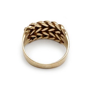 K&ouml;nnte beinhalten: Ein goldener Ring mit einem geflochtenen Design. Der Ring besteht aus einem dicken Metallband und hat eine glatte Oberfl&auml;che.