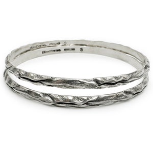 May include: Two silver bangles with a textured, wavy design. The bangles are stacked on top of each other and have the words "sterling" and "3" engraved on the inside.