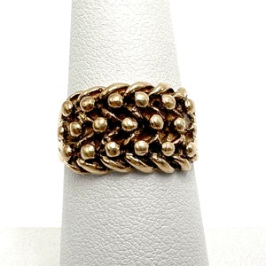 K&ouml;nnte beinhalten: Ein goldener Ring mit einem strukturierten Design aus miteinander verbundenen Ketten und kleinen Perlen.