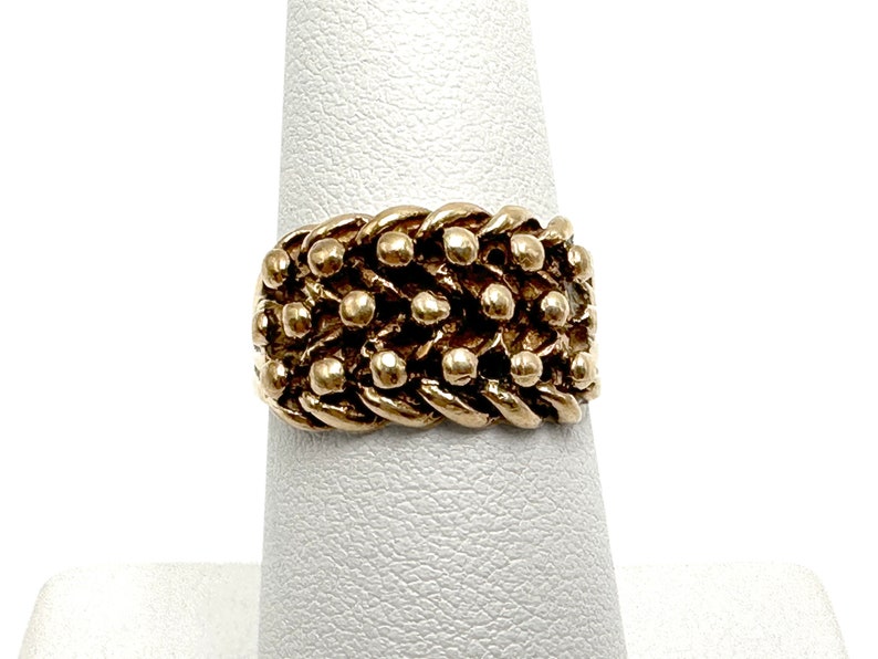 K&ouml;nnte beinhalten: Ein goldener Ring mit einem strukturierten Design aus miteinander verbundenen Ketten und kleinen Perlen.