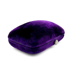 May include: A plush, deep purple velvet jewelry box with a rounded shape. The box has a small, off-white clasp on the side. The velvet material gives the box a luxurious appearance.