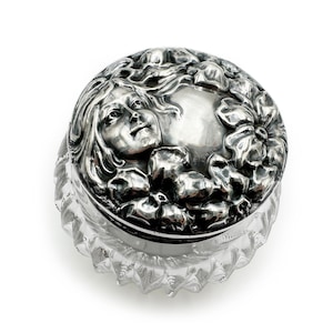 May include: A silver-plated glass jar with a lid featuring a raised design of a woman's face surrounded by flowers.