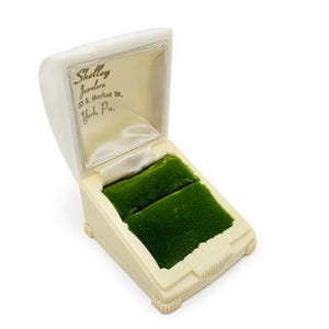 May include: An open, off-white jewelry box with a green velvet interior. The box has a hinged lid and is labeled "Shelley Jewelers, 53 E. Market St., York, Pa." The box is designed to hold rings or small jewelry items.
