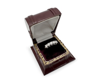 Op de afbeelding: Een diamanten ring met vijf stenen rust in een open, antieke, bordeauxrode leren sieradendoos. De doos heeft een goudkleurige rand en een donker bordeauxrood fluwelen interieur. De ring is het middelpunt, tegen de donkere fluweel.