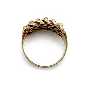 K&ouml;nnte beinhalten: Ein goldener Ring mit geflochtenem Design. Der Ring besteht aus einer Reihe von ineinandergreifenden Schlaufen, die eine strukturierte Oberfl&auml;che erzeugen.