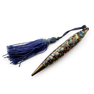 May include: A patterned pen with a dark blue tassel. The pen's barrel features a colorful design, and it has a bronze-coloured tip. The tassel is made of dark blue threads and is attached to the pen.