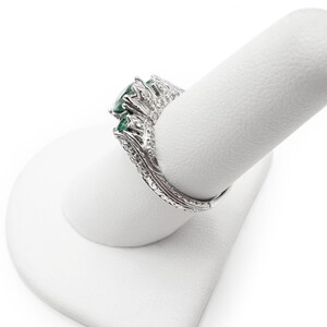 May include: A silver ring with a large emerald green gemstone surrounded by smaller diamonds. The ring is on a white ring display stand.
