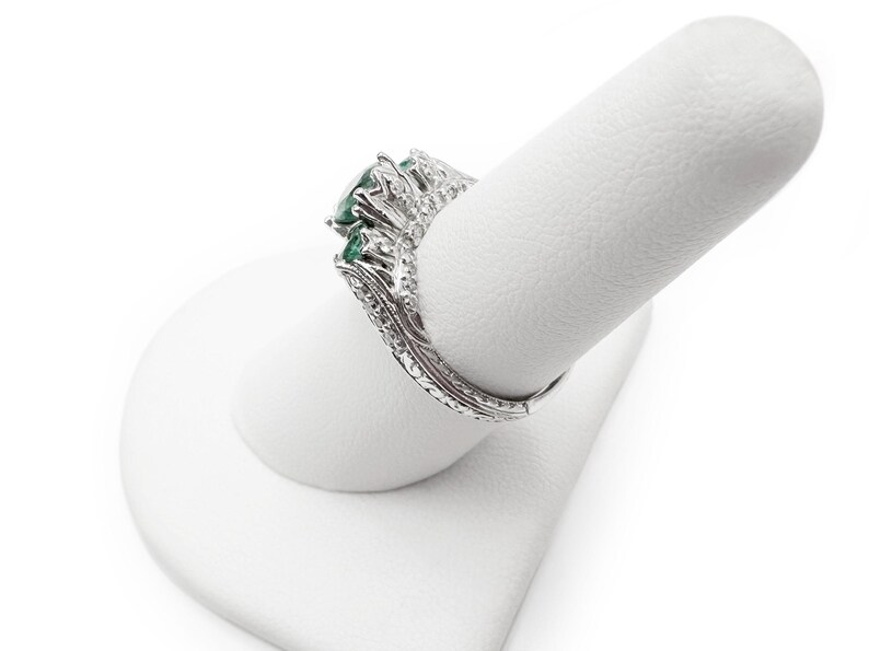 May include: A silver ring with a large emerald green gemstone surrounded by smaller diamonds. The ring is on a white ring display stand.