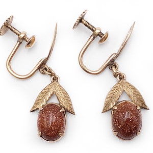 May include: A pair of gold tone clip-on earrings with brown stone accents. Each earring features a teardrop-shaped stone with a gold tone leaf design.